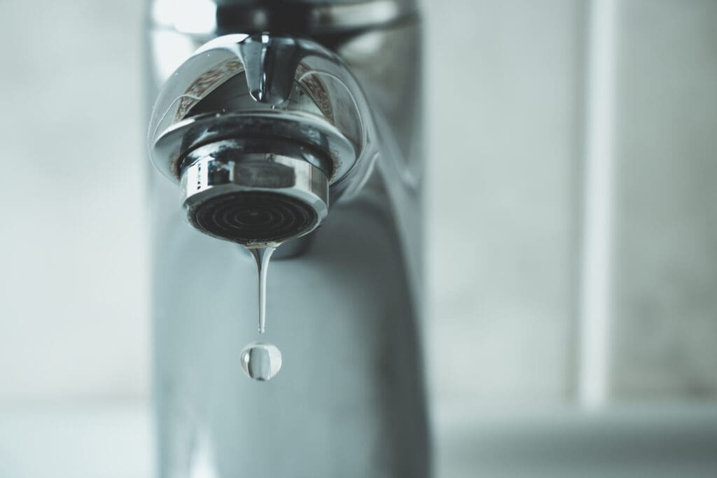 Close-up of a metal faucet with a droplet of water about to fall from its spout, against a blurred, light-colored background.