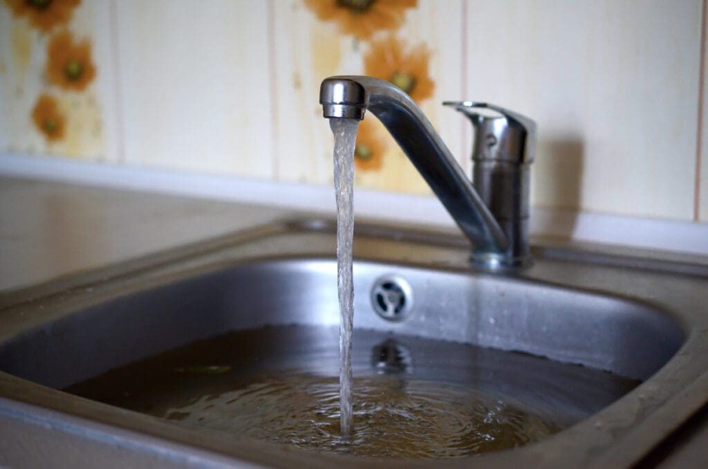 A modern metal kitchen faucet releases a stream of water into a stainless steel sink. The sink is partially filled with murky, brownish water. The background features yellow flower-patterned wallpaper.