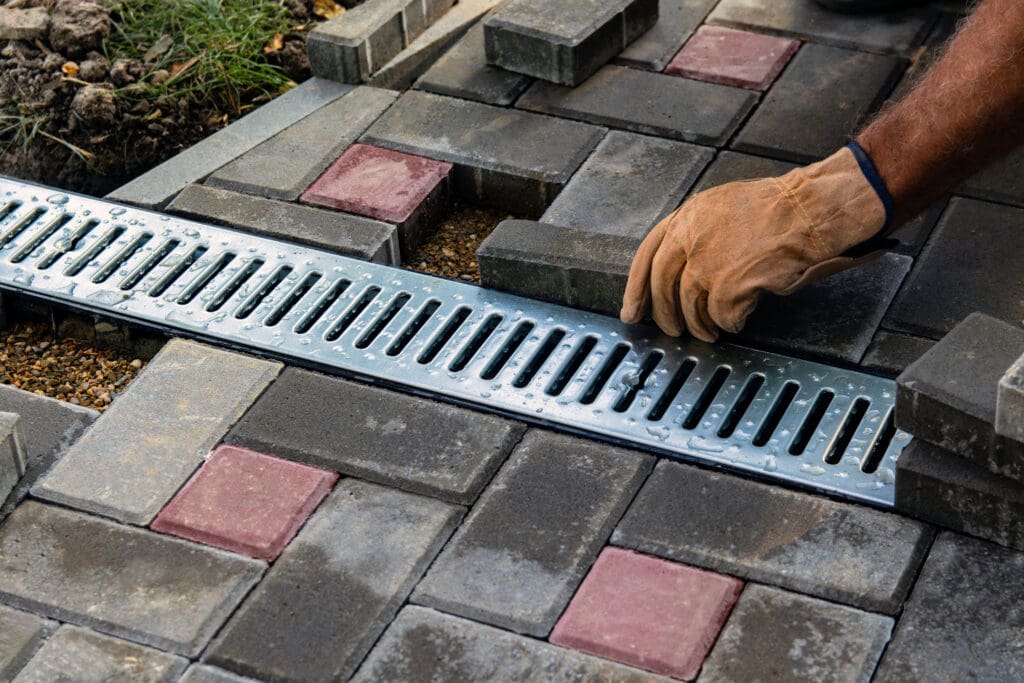 A person wearing a work glove is installing interlocking paving stones around a metal drainage grate in an outdoor area. The pavers are gray with some red accents.
