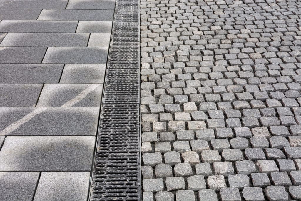 Close-up of a paved surface showing two different patterns: large rectangular gray tiles on the left and small square cobblestones on the right, separated by a metal drainage grate running vertically.