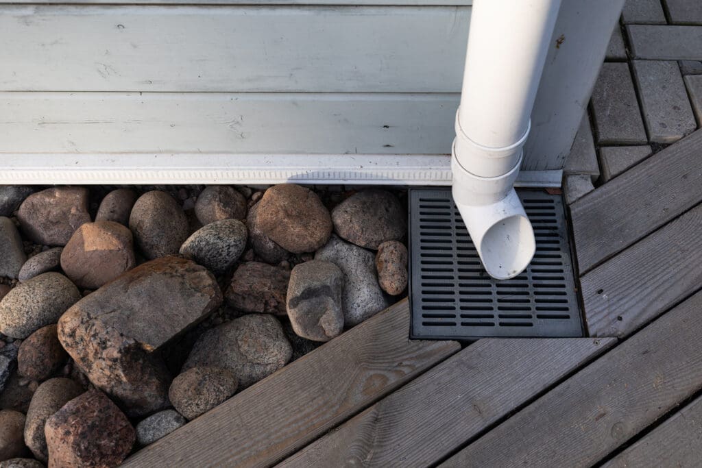 A white downspout from a building drains into a black grate set in a wooden deck next to a section of rocks and gray paving stones.