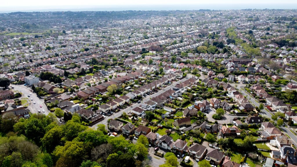 Aerial view of a sprawling suburban neighborhood with rows of houses, tree-lined streets, and green spaces, extending toward the horizon under a clear sky.