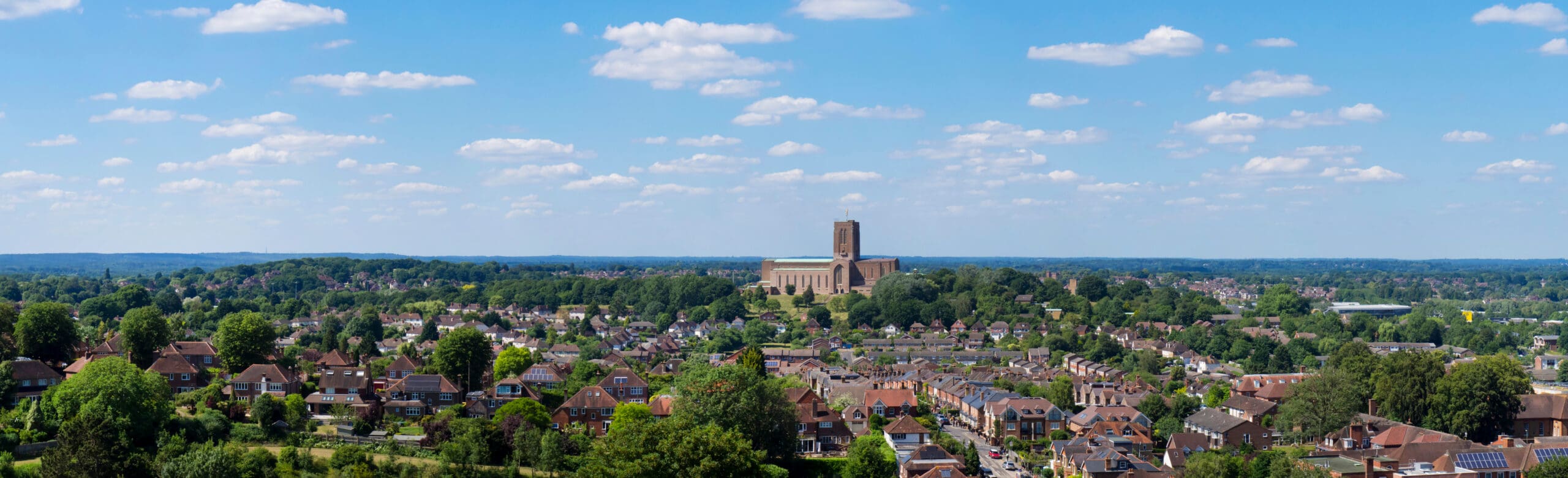 A panoramic view of a town with many houses surrounded by greenery, with a large church or cathedral prominently in the center, under a blue sky with scattered clouds.