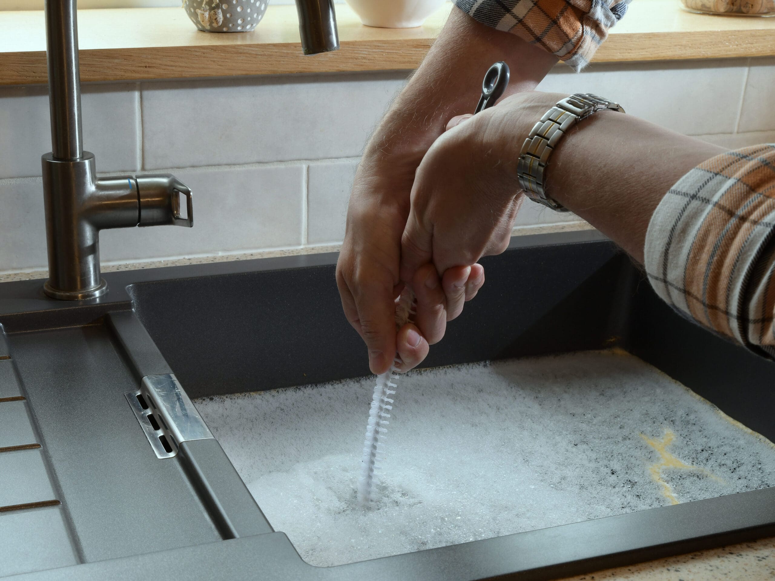 A person wearing a plaid shirt cleans a dish brush under running water in a kitchen sink filled with soapy water. The countertop is light-colored, and the faucet is modern.