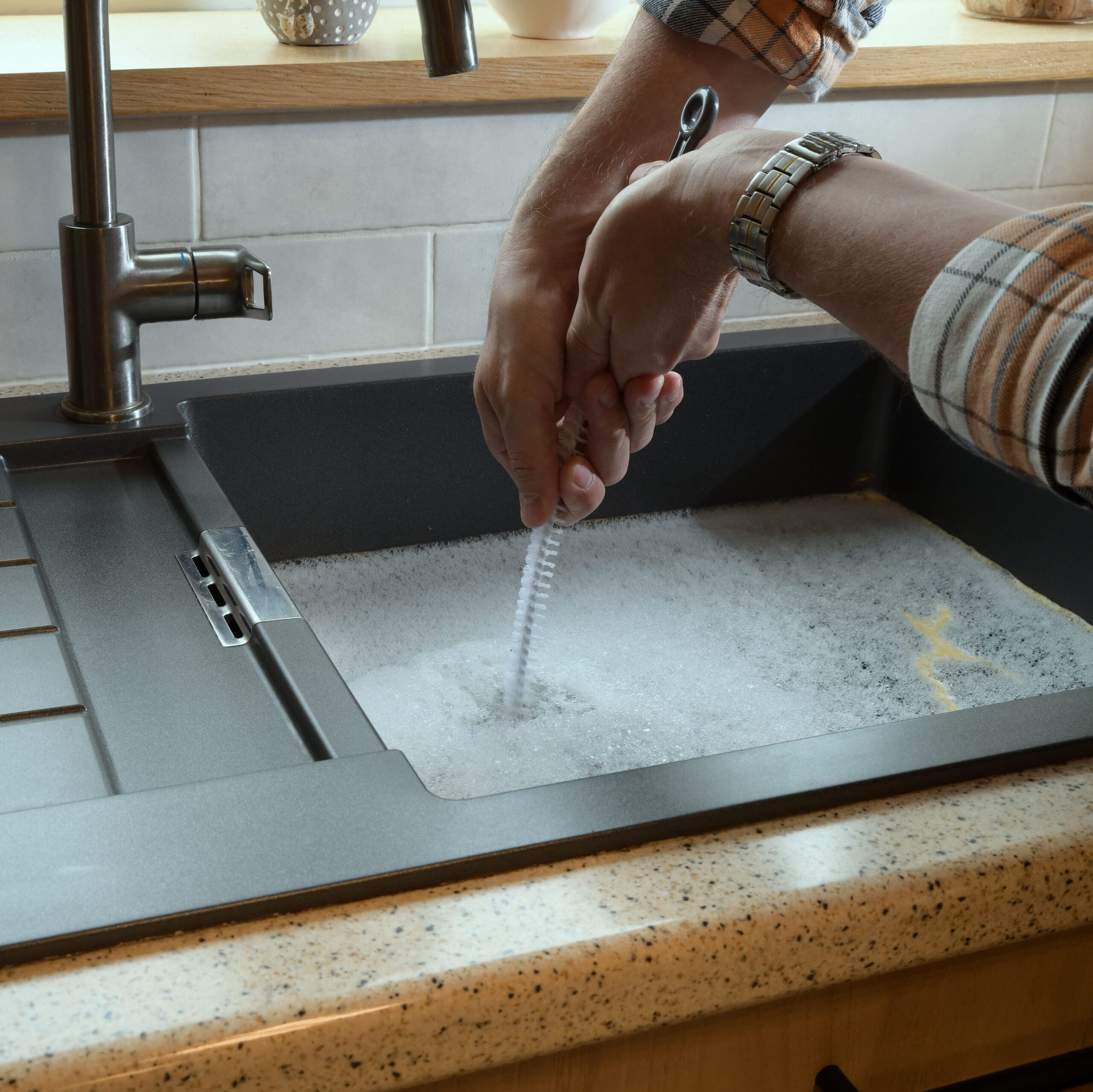 A person wearing a plaid shirt cleans a dish brush under running water in a kitchen sink filled with soapy water. The countertop is light-colored, and the faucet is modern.