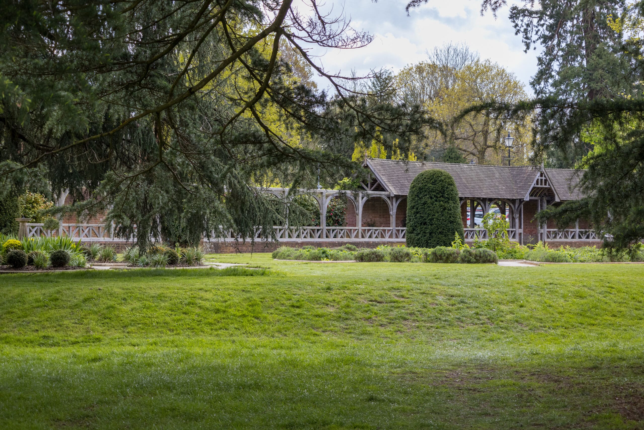 A covered wooden walkway with lattice railings stands amid lush green grass and trees, with branches partially framing the view and bushes and plants surrounding the structure in a park setting.