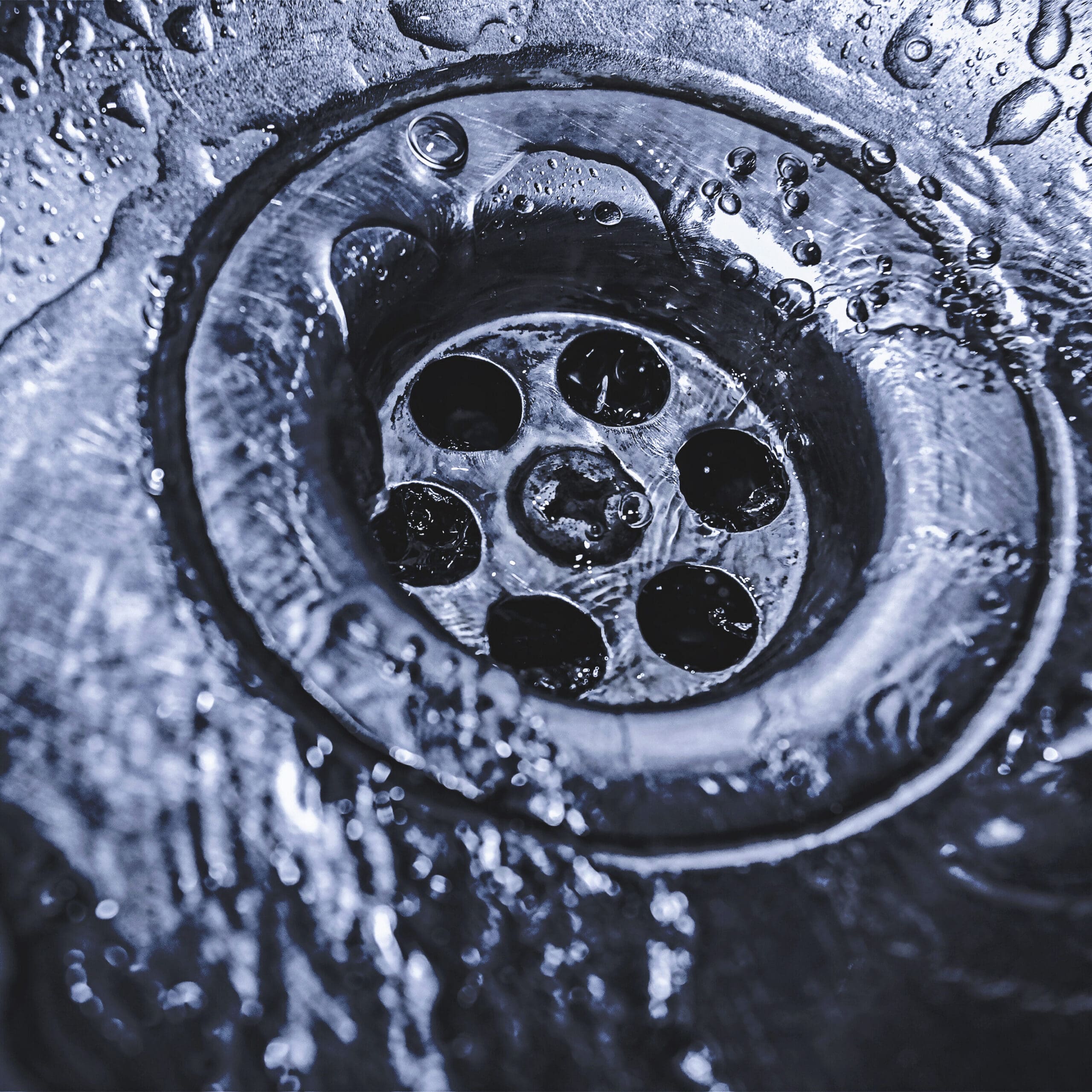 Close-up of water flowing into a metal sink drain, with water droplets and reflections visible on the surface around the drain.