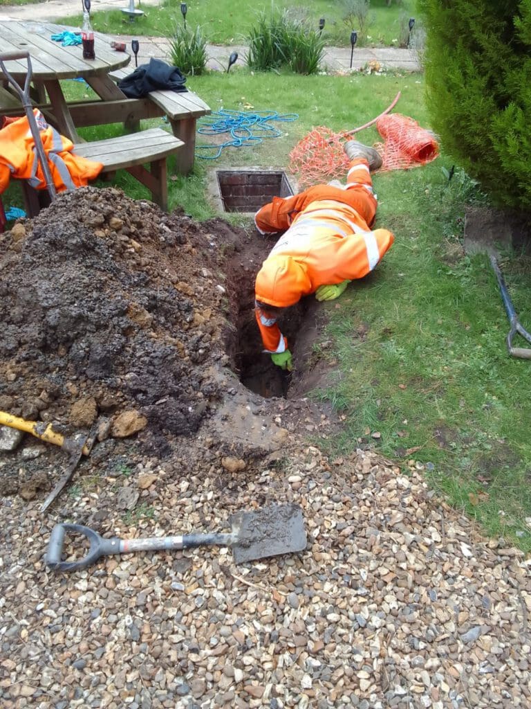 A worker in an orange safety suit is lying on the ground, reaching into a narrow trench next to a pile of dirt and a gravel path. A shovel and tools are nearby, with grass and garden furniture in the background.