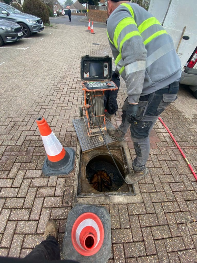 A worker in a hi-vis jacket operates a drain inspection camera over an open manhole in a paved parking area, with two orange traffic cones nearby for safety.