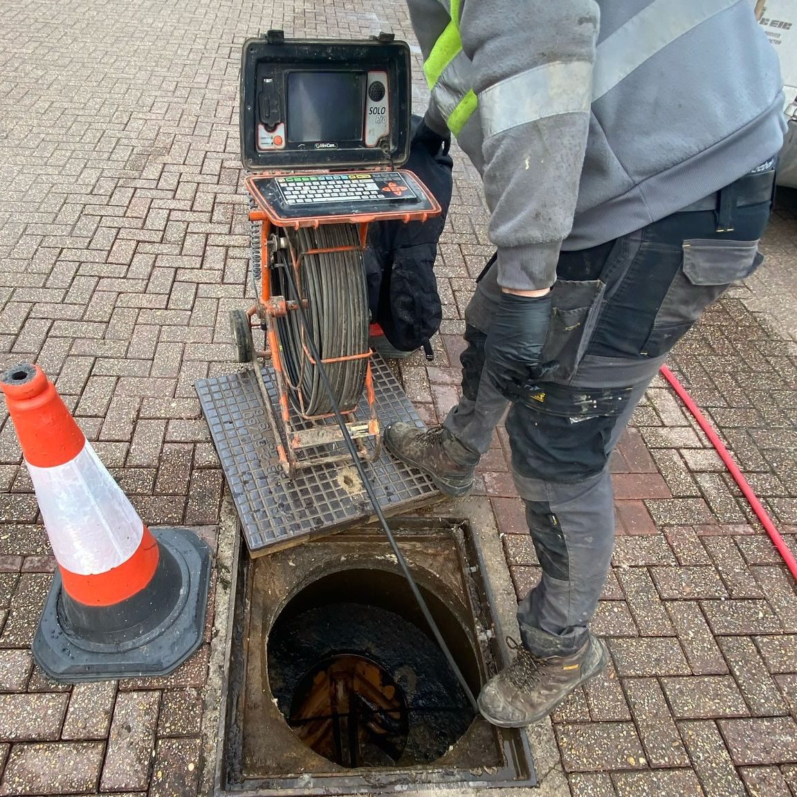 A worker in a hi-vis jacket operates a drain inspection camera over an open manhole in a paved parking area, with two orange traffic cones nearby for safety.