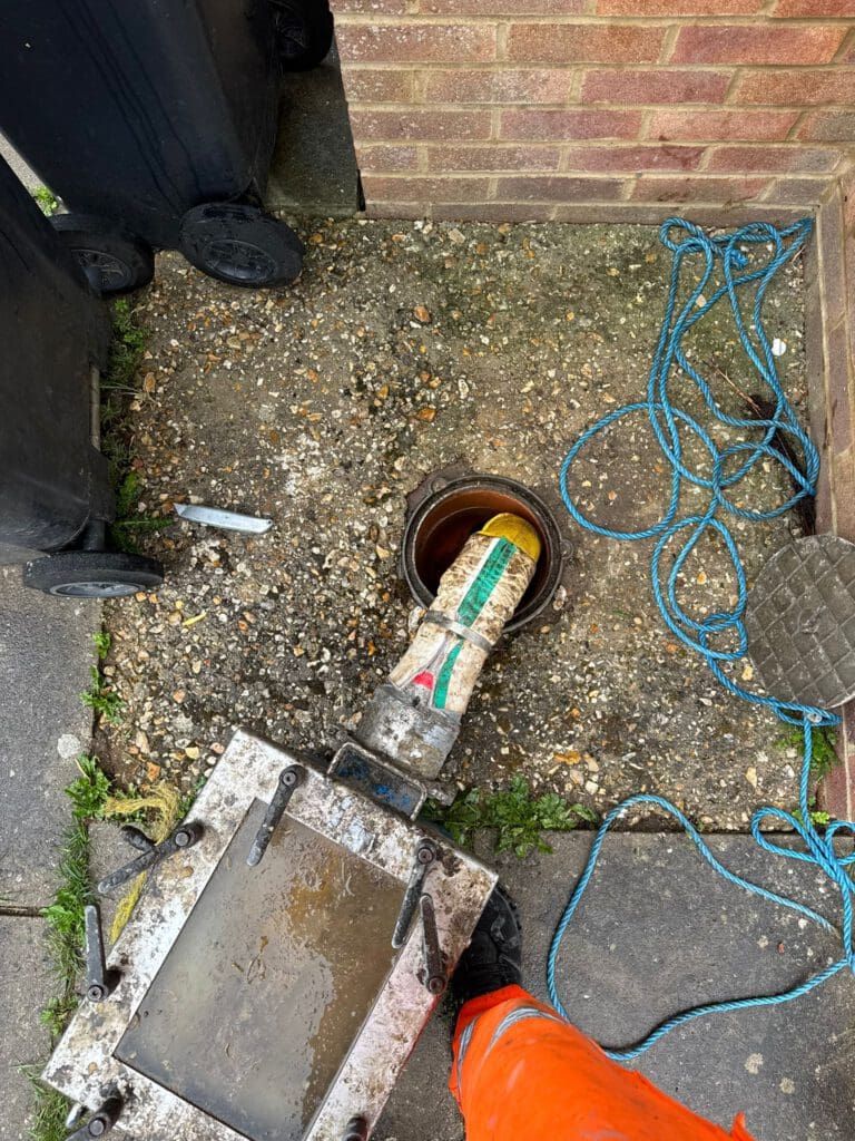 A person in orange work clothes uses equipment to clean or inspect a drain on a concrete area next to a brick wall. Blue rope, two black bins, and a metal tool are nearby. A hose extends into the drain opening.