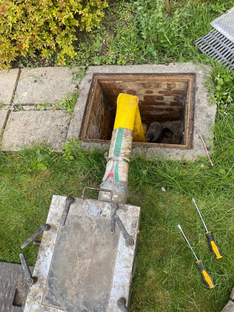 A yellow pipe is being inserted into an open brick-lined manhole in a grassy area, with paving stones nearby. Tools, including screwdrivers, lie on the grass beside the manhole cover.