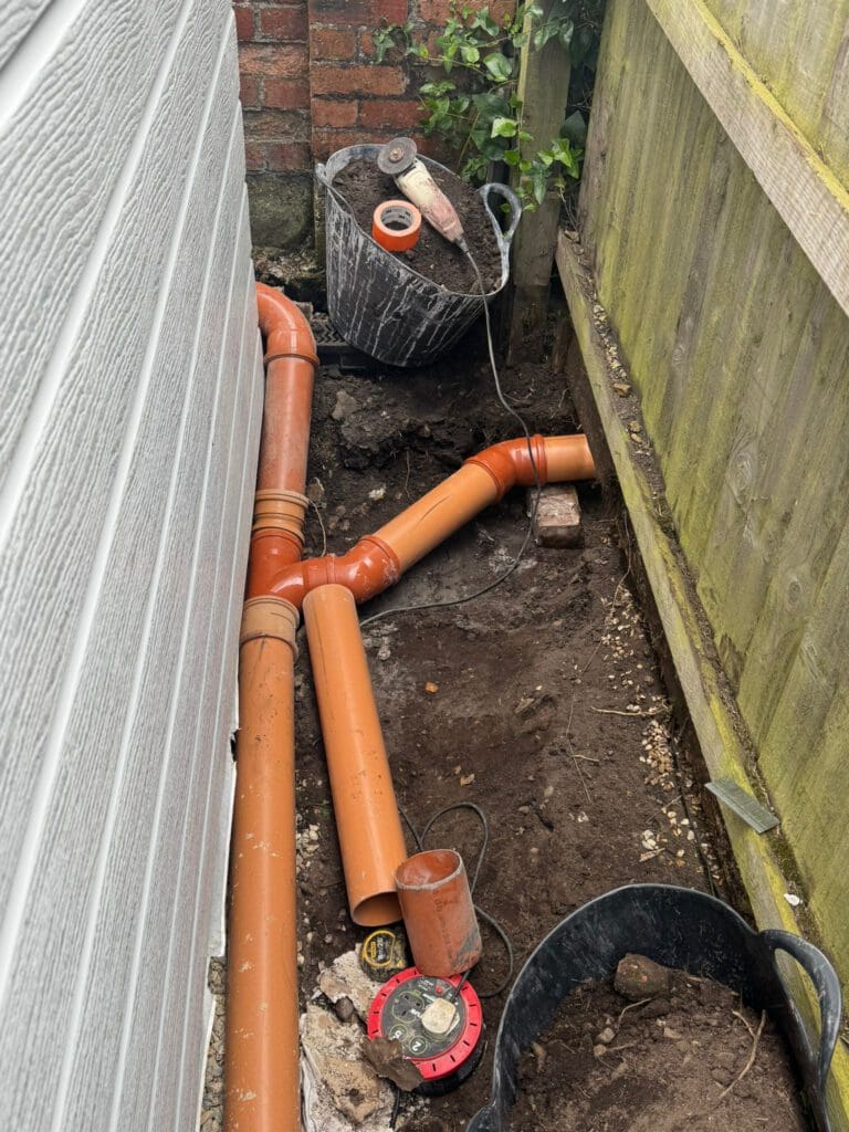Orange PVC drainage pipes being installed in a narrow space between a house wall and a wooden fence. Construction tools and a black bucket with rubble are nearby on the soil.