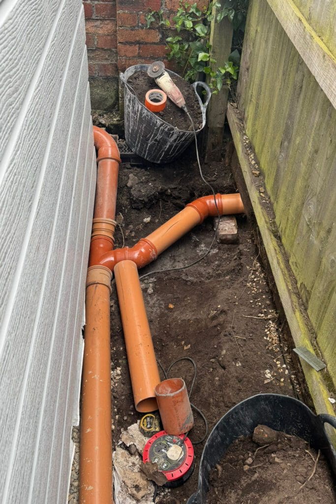 Orange PVC drainage pipes being installed in a narrow space between a house wall and a wooden fence. Construction tools and a black bucket with rubble are nearby on the soil.