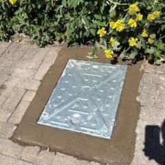 A metal utility cover is set in a rectangular patch of fresh concrete surrounded by paving stones. Yellow flowers and green plants border the top of the image. A shadow is visible in the lower right corner.
