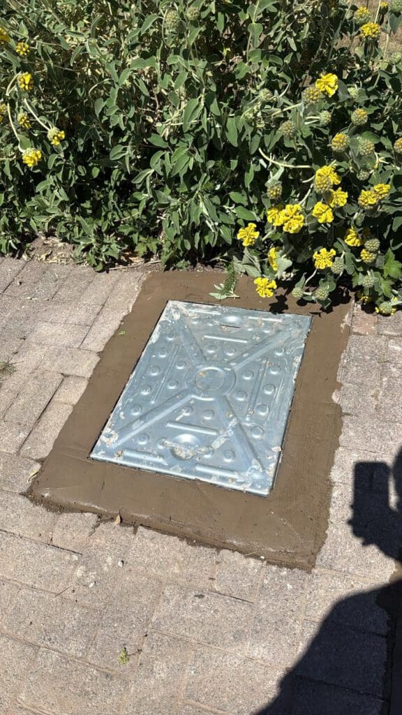 A metal utility cover is set in a rectangular patch of fresh concrete surrounded by paving stones. Yellow flowers and green plants border the top of the image. A shadow is visible in the lower right corner.