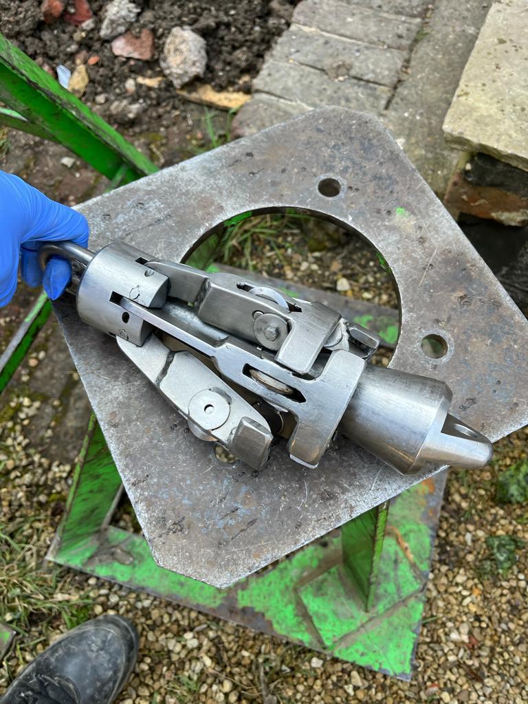 A person wearing a blue glove holds a complex, metallic mechanical tool with moving parts, placed on a weathered metal surface outdoors near gravel and bricks.