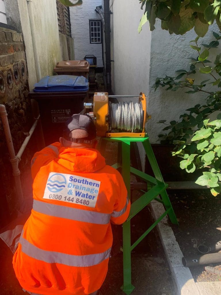 A person in a bright orange "Southern Drainage & Water" jacket operates a cable winch next to bins and brick walls in a narrow outdoor alleyway with sunlight and nearby greenery.