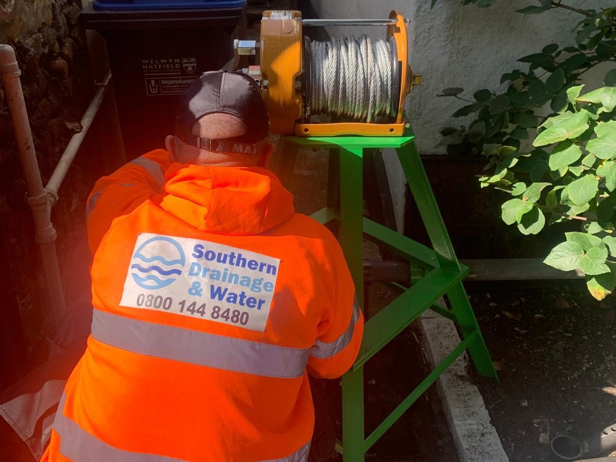 A person in a bright orange "Southern Drainage & Water" jacket operates a cable winch next to bins and brick walls in a narrow outdoor alleyway with sunlight and nearby greenery.