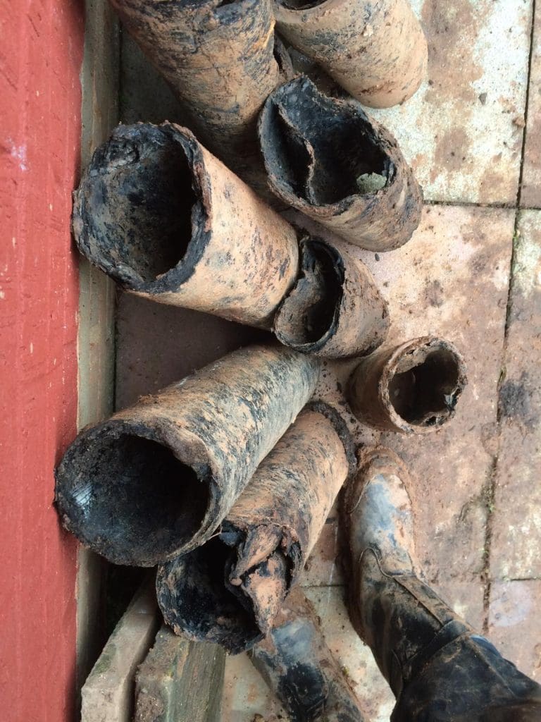Several old, heavily rusted and corroded metal pipes are stacked on a tiled ground near a red wall. A person wearing muddy boots stands next to the pipes.