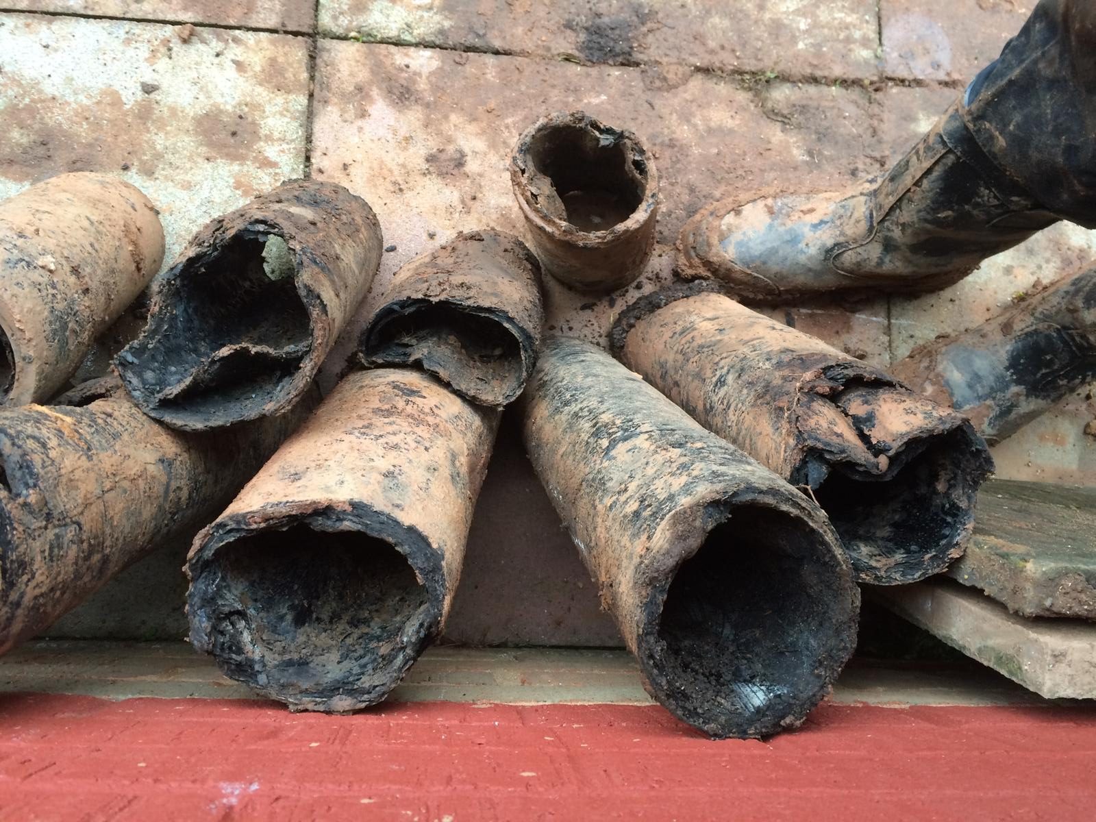 Several old, heavily rusted and corroded metal pipes are stacked on a tiled ground near a red wall. A person wearing muddy boots stands next to the pipes.