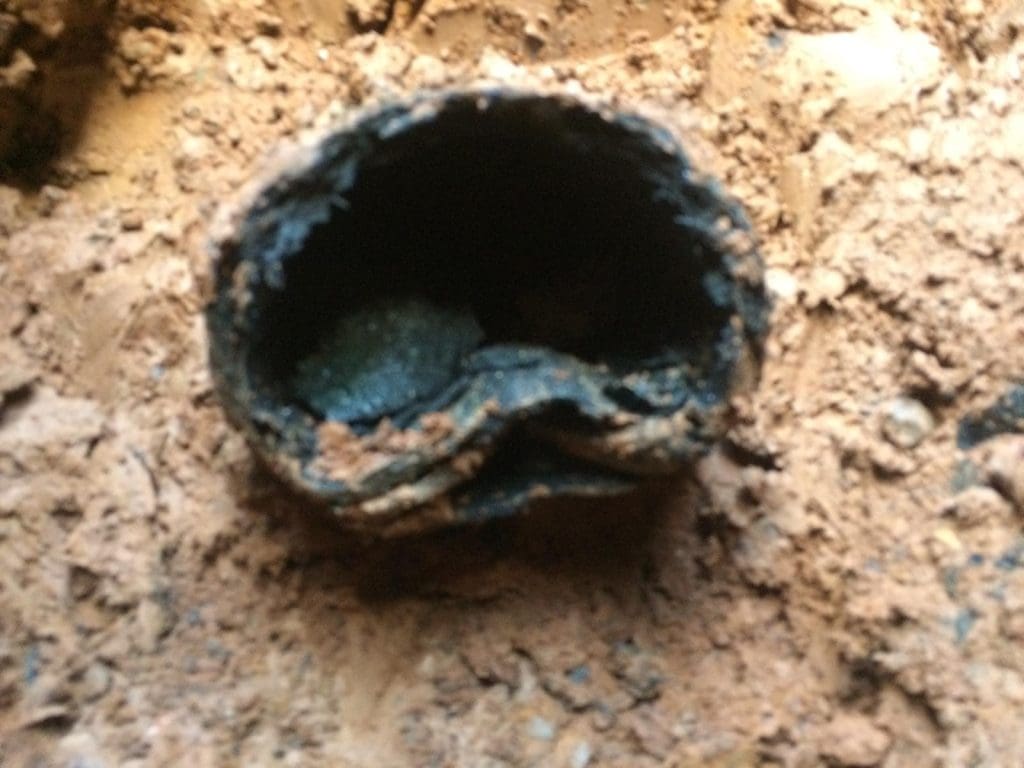 A close-up of a broken, corroded metal pipe surrounded by brown dirt. The pipe is partially collapsed and clogged, with muddy debris visible both inside and around the opening.