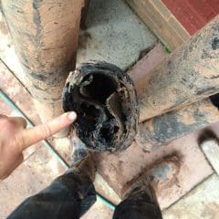 A person points at the inside of a clogged, mud-covered pipe filled with compacted debris. The person is standing on a dirt-stained surface with other dirty pipes and their muddy boots visible nearby.