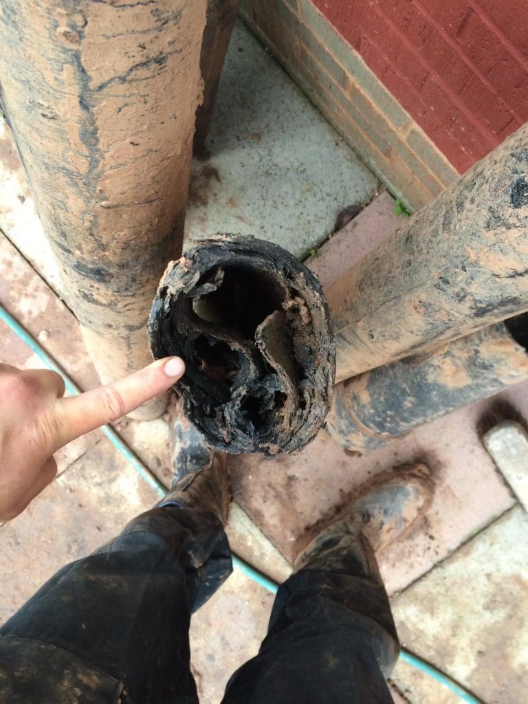 A person points at the inside of a clogged, mud-covered pipe filled with compacted debris. The person is standing on a dirt-stained surface with other dirty pipes and their muddy boots visible nearby.