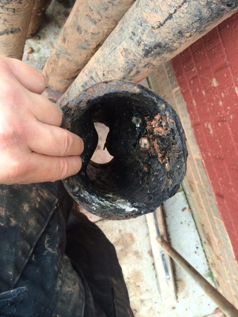 A hand holds a heavily clogged, corroded pipe with significant buildup inside. The pipe is dirty and surrounded by mud and debris, next to a red brick wall.