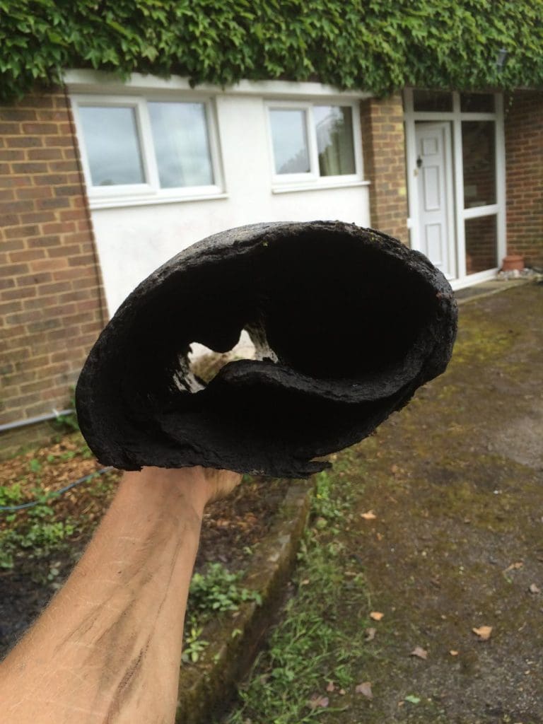 A person holds up a large, blackened, hollow cylinder that appears to be a severely burnt or clogged pipe, with a residential house and garden visible in the background.