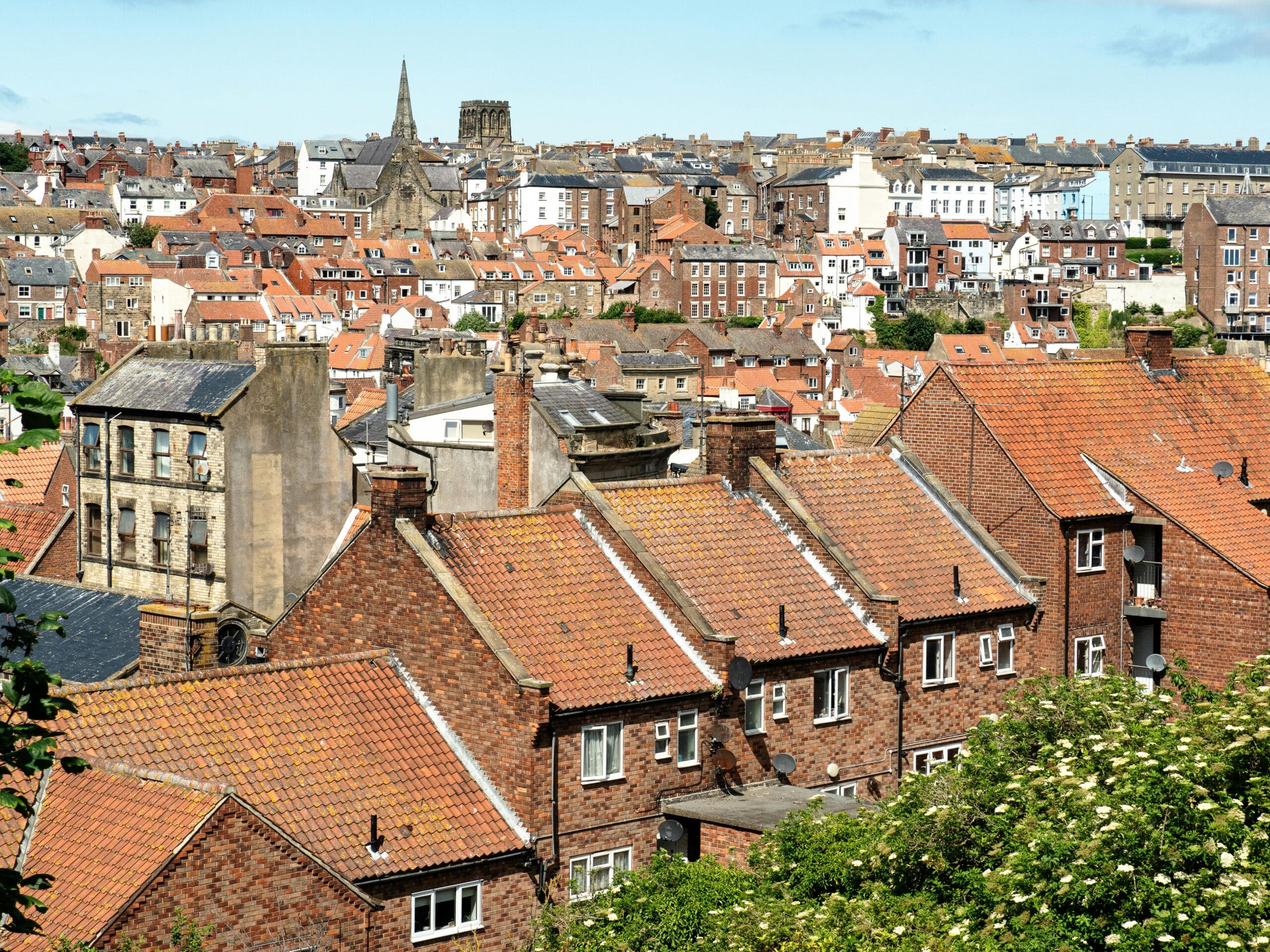A scenic view of a historic town with rows of red-roofed brick houses, old buildings, and church spires under a bright blue sky. Greenery frames the foreground.